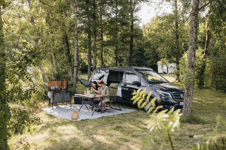 Paar isst draußen am Mercedes Vito Campervan auf einem Waldstellplatz mit Hängematte und mobiler Outdoor-Küche